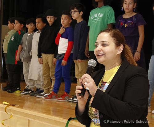 A woman speaks into a microphone with students standing on a stage behind her.